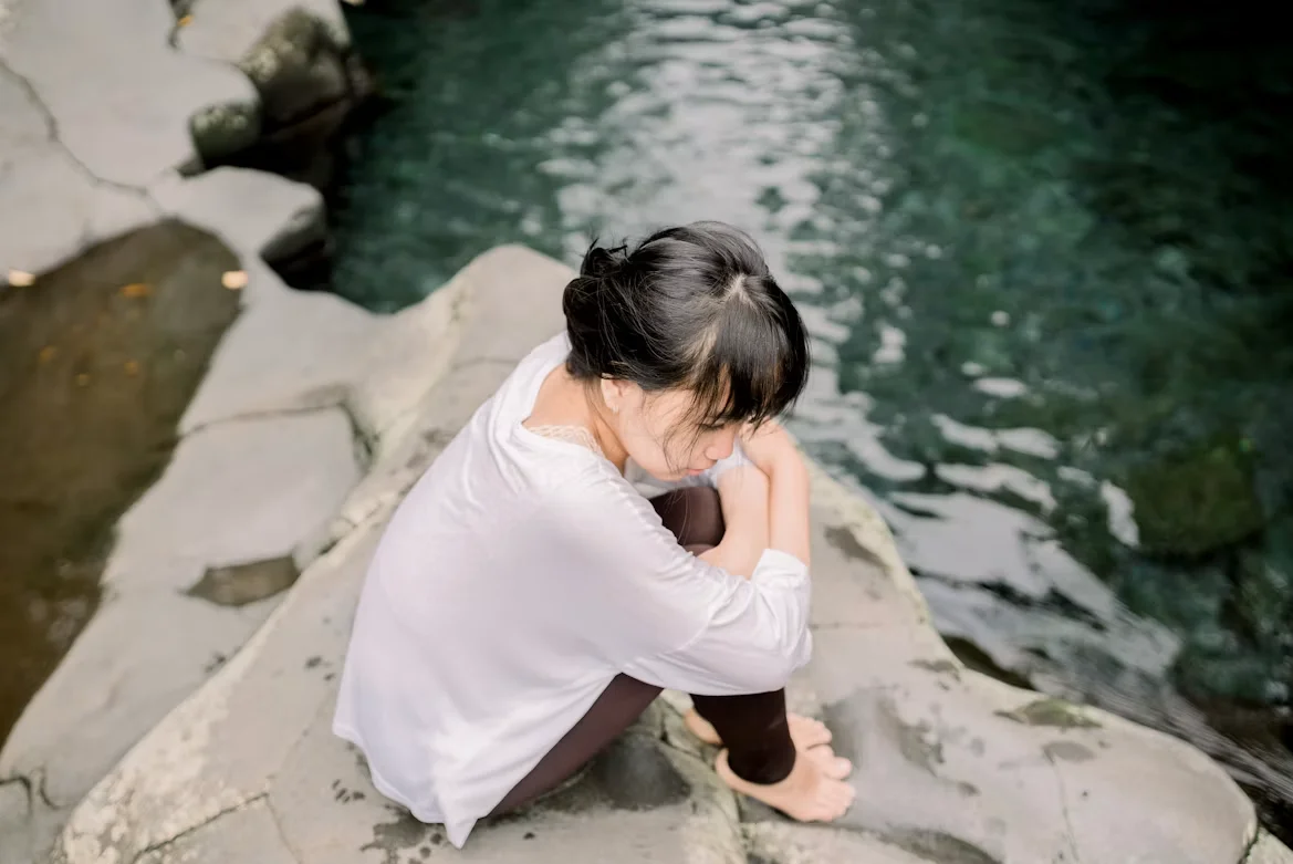 A woman sitting alone on rocks beside a calm pool, reflecting on her health